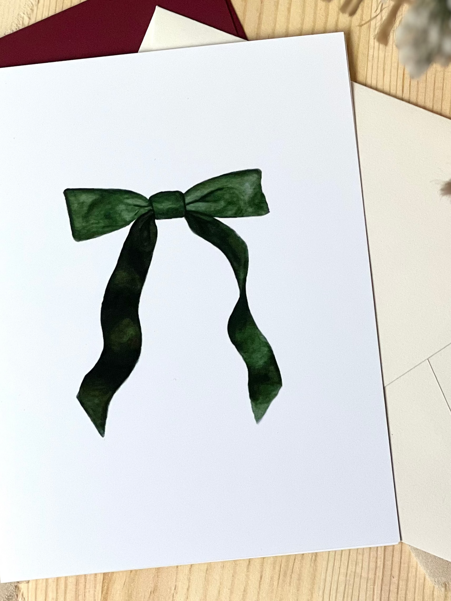 A greeting card with a green ribbon design on a white background, displayed on a wooden surface with an envelope and decorative material in the background.