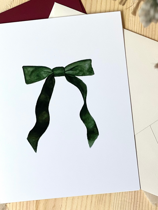 A greeting card with a green ribbon design on a white background, displayed on a wooden surface with an envelope and decorative material in the background.
