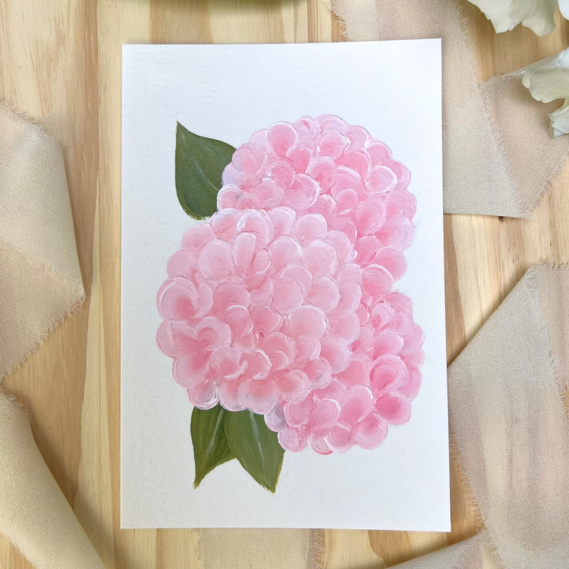 A square white art print featuring a painted pink hydrangea with green leaves, styled on a light wood surface with beige fabric accents.