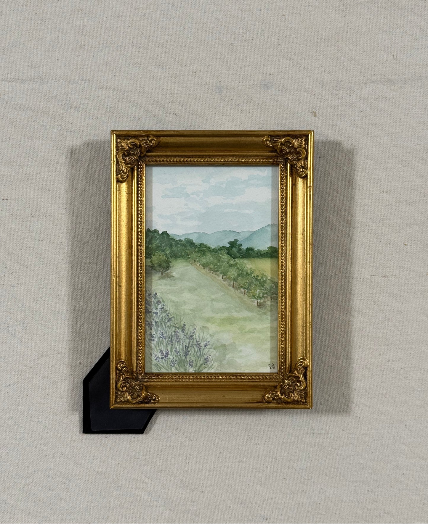 A framed landscape painting featuring a lavender patch ,an apple orchard, and distant mountains, housed in an ornate gold frame