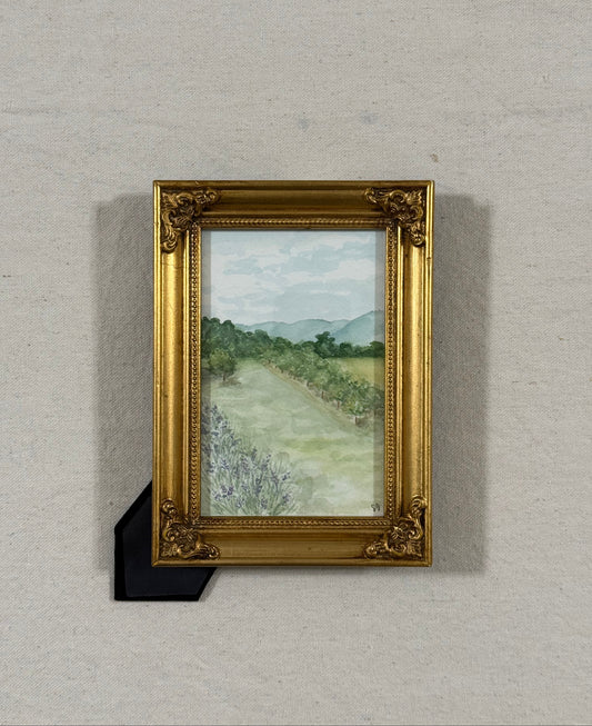 A framed landscape painting featuring a lavender patch ,an apple orchard, and distant mountains, housed in an ornate gold frame