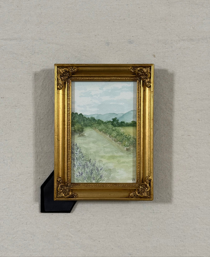A framed landscape painting featuring a lavender patch ,an apple orchard, and distant mountains, housed in an ornate gold frame